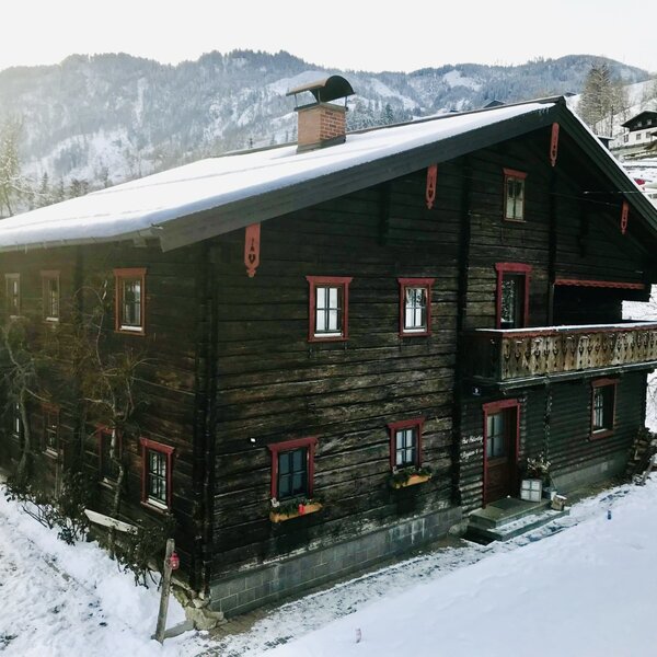 The dark wooden farmhouse exterior with red window frames, a balcony, and a snow-covered roof, set against a backdrop of snowy mountains.