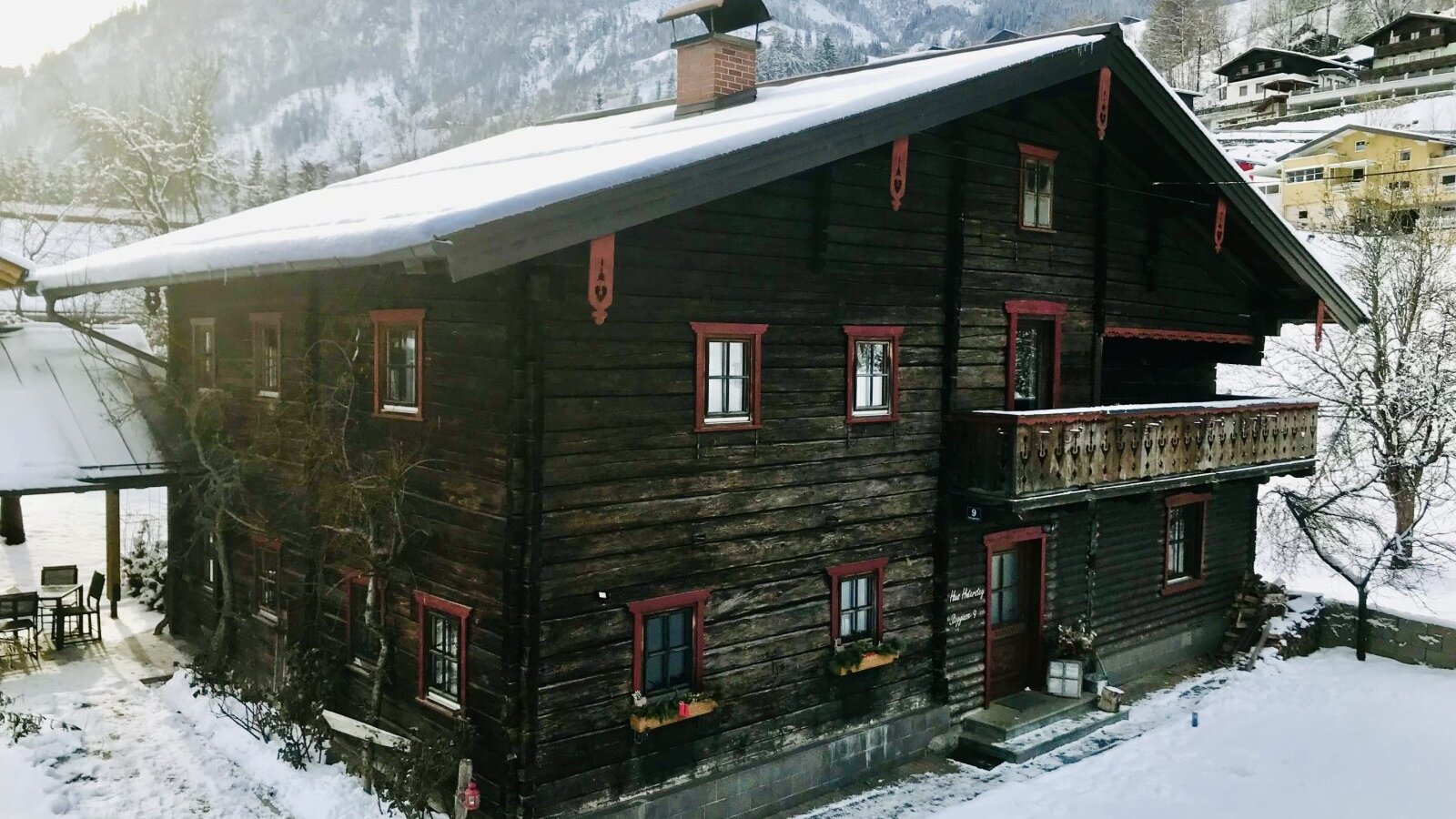 The dark wooden farmhouse exterior with red window frames, a balcony, and a snow-covered roof, set against a backdrop of snowy mountains.