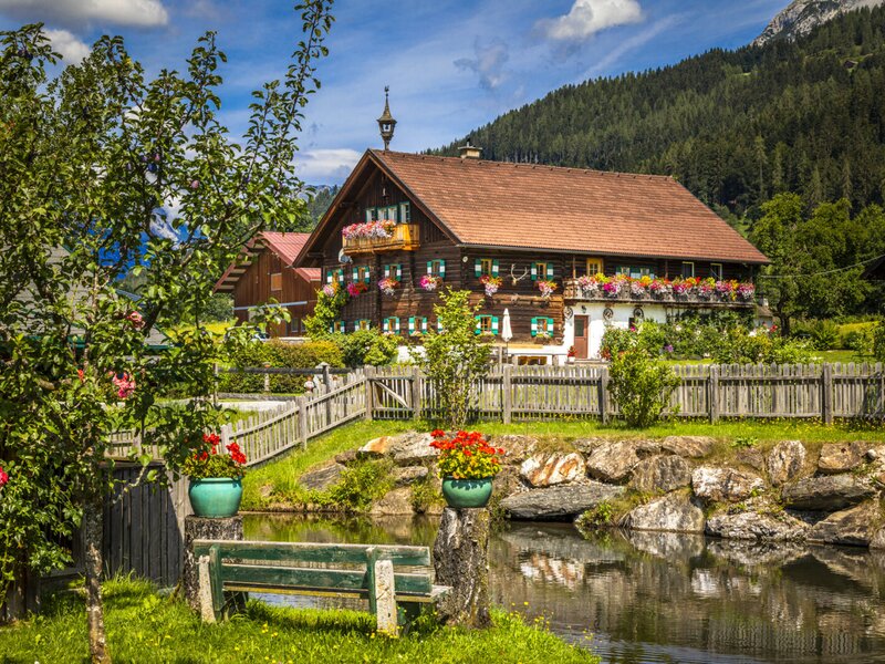 The farmhouse exterior, featuring a wooden facade, flower-filled balconies, and green shutters, is situated next to a small pond with a bench and surrounded by lush greenery and mountains.
