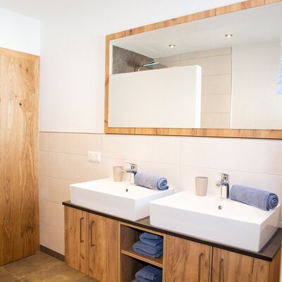 The farm house bathroom features double rectangular sinks on a wooden vanity, a large mirror, a towel warmer, and a shower area visible in the mirror's reflection.