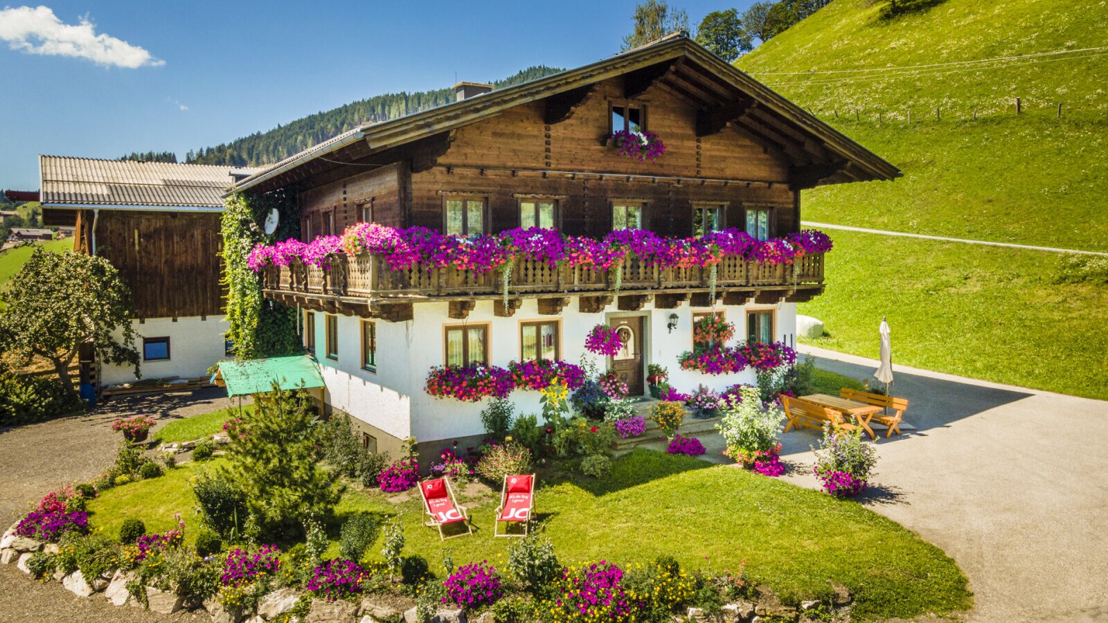 The farmhouse exterior features traditional wooden balconies and window boxes overflowing with purple flowers, a front garden with red deck chairs, and an outdoor dining set.