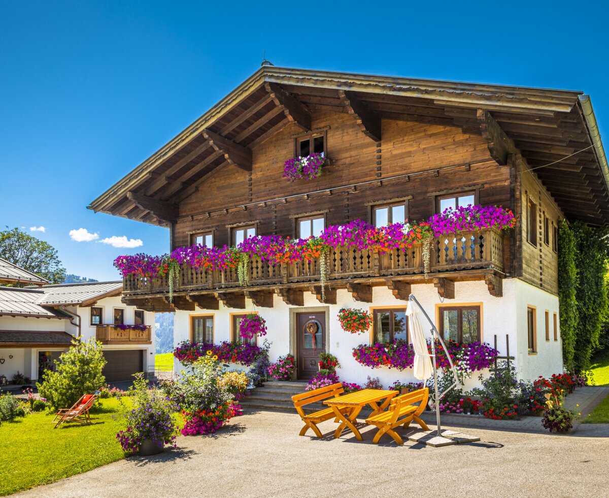 The farm house exterior features wooden balconies adorned with colorful flowers and an outdoor seating area with a wooden table and benches.