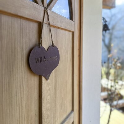 The entrance to the farmhouse, marked by a wooden door with a heart-shaped "Willkommen" sign.