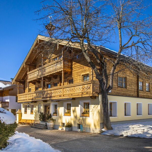 The exterior of the Farm House, featuring traditional wooden architecture, multiple carved balconies, and a paved entrance in winter.