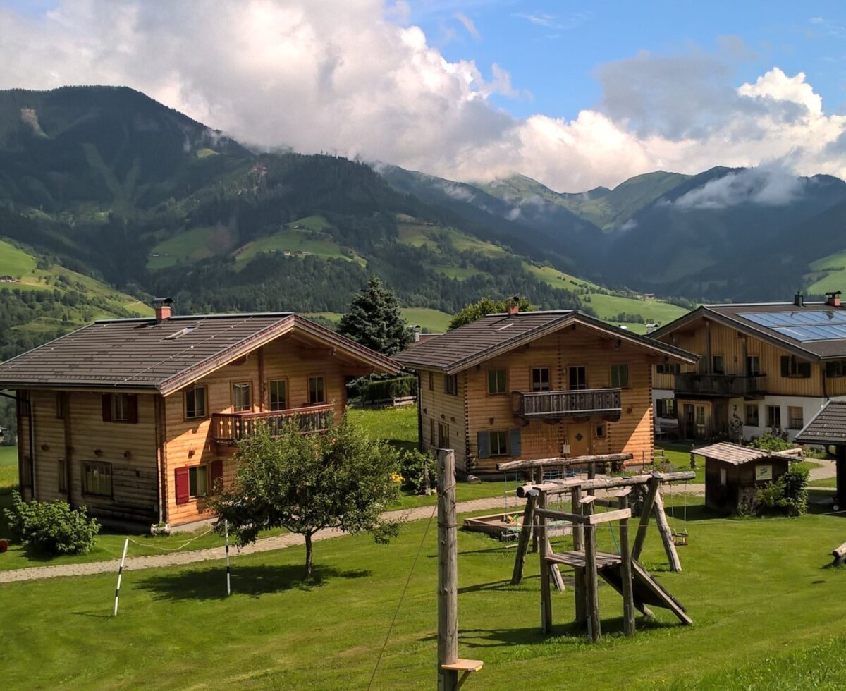 The farm houses are surrounded by a green lawn with a children's playground, set against a backdrop of mountains.