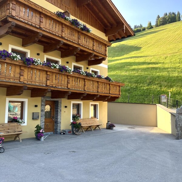 The farm house features a yellow exterior, multiple wooden balconies adorned with flower boxes, and a paved courtyard with benches, set against a green hillside.