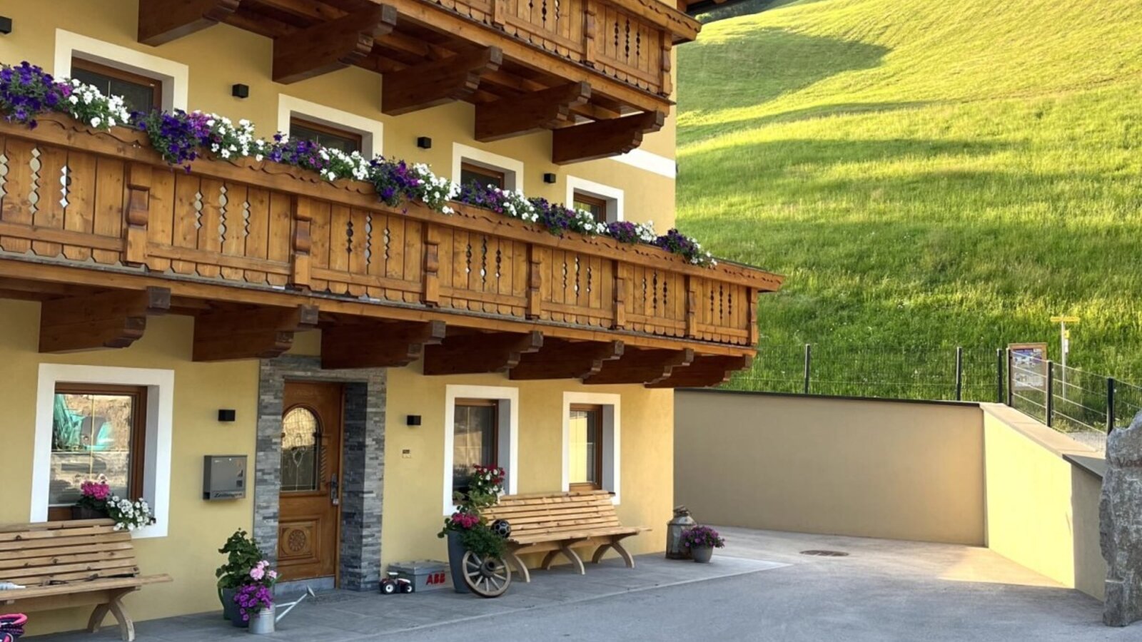 The farm house features a yellow exterior, multiple wooden balconies adorned with flower boxes, and a paved courtyard with benches, set against a green hillside.