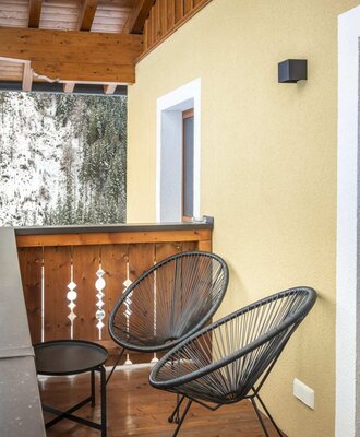 Balcony of the farmhouse with two chairs, a table, and a view of snow-covered trees.