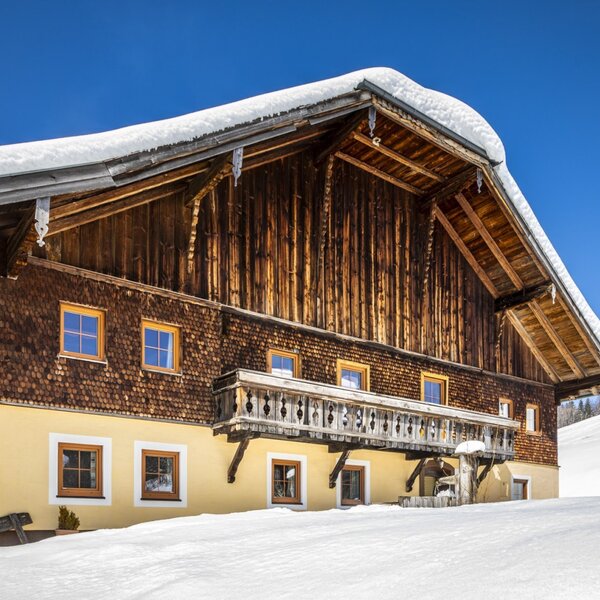 The farm house exterior, featuring a traditional wooden upper facade, a yellow lower level, and a balcony, surrounded by snow with ski lifts visible in the background.