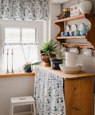 Kitchen area in the Bed and Breakfast with a window, floral curtains, a wooden shelf, and dishes.