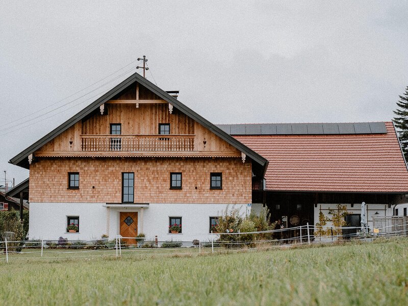 The traditional wooden facade of the restored farmhouse, featuring a balcony and an adjacent building section.