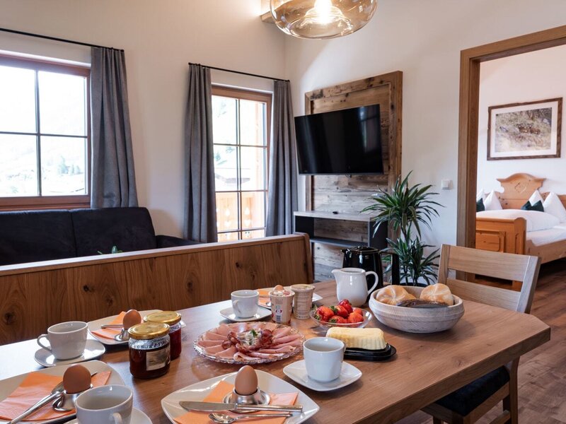 The dining area in the Bauernhof is set for breakfast, offering a view of the living area with a sofa and TV, and an adjoining bedroom.