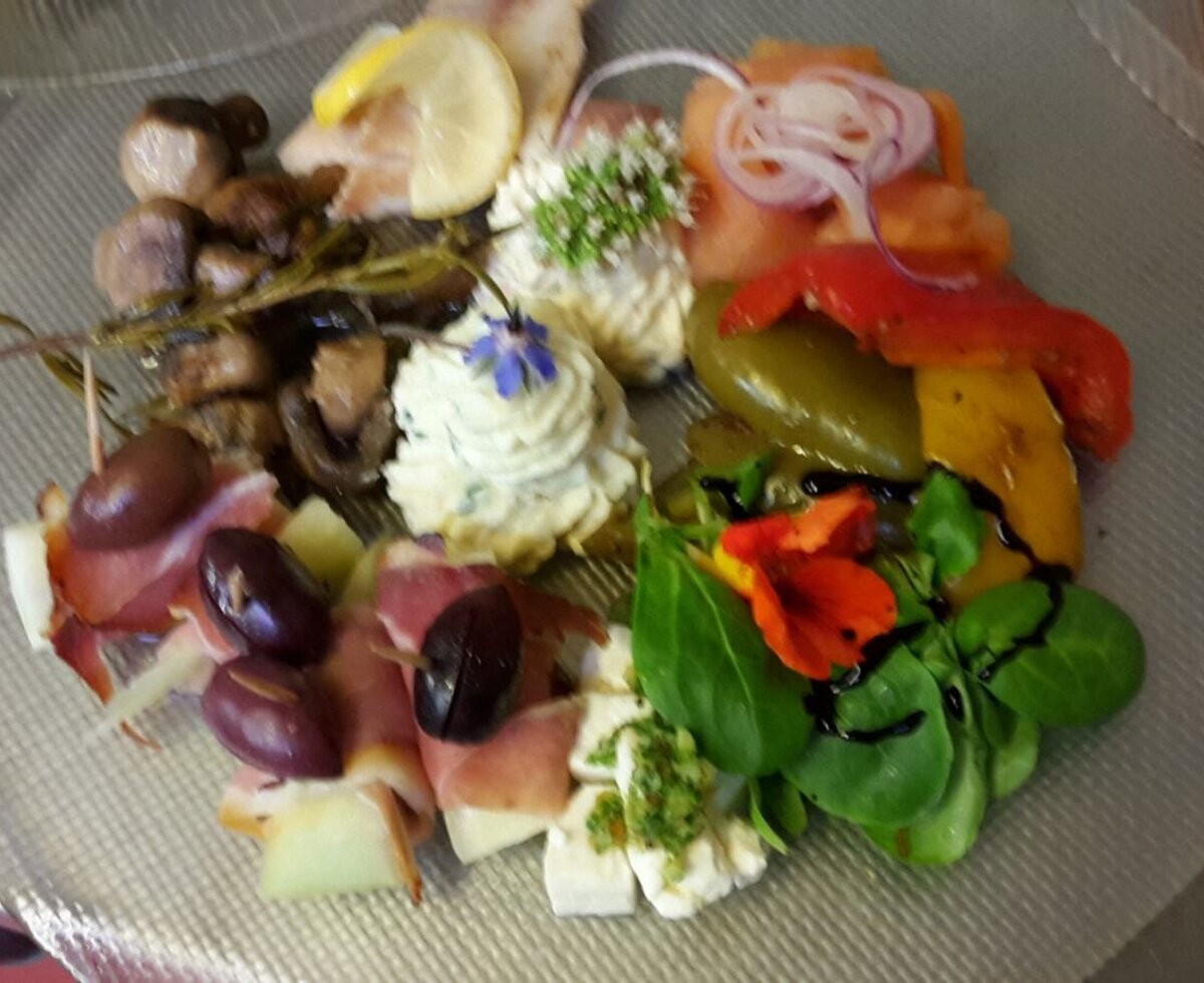 A mixed appetizer plate featuring cured meats, olives, cheeses, marinated mushrooms, bell peppers, and garnished dips.