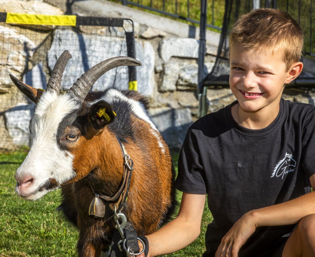 A smiling boy with a friendly goat on the grassy grounds of the family-friendly farmhouse.