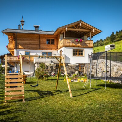 The farmhouse's inviting garden featuring a swingset, a trampoline, and a spacious green lawn set against a scenic hillside.