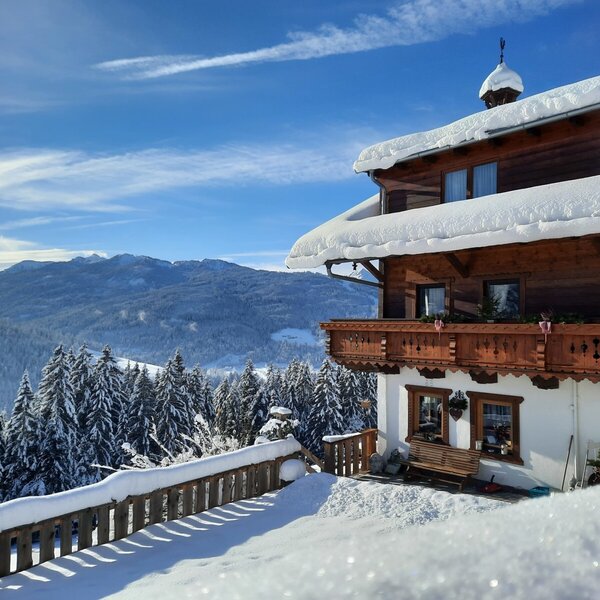 The snow-covered farm house features a traditional wooden balcony and views of the surrounding mountains and forests.