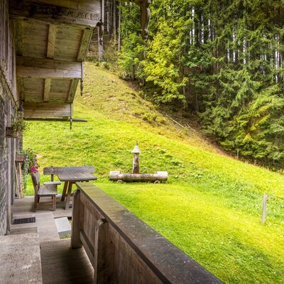 The mountain hut's rustic wooden porch, featuring a cozy outdoor seating area beside a lush green slope and dense pine forest.