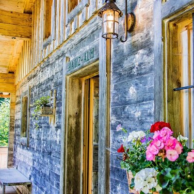 The farmhouse exterior features rustic wooden walls, a traditional lantern, colorful window boxes, and a wooden balcony with a bench.