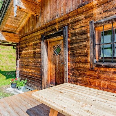 The wooden exterior of the farmhouse features a deck with a picnic table, an entry door, and a traditional window, overlooking a green hillside.