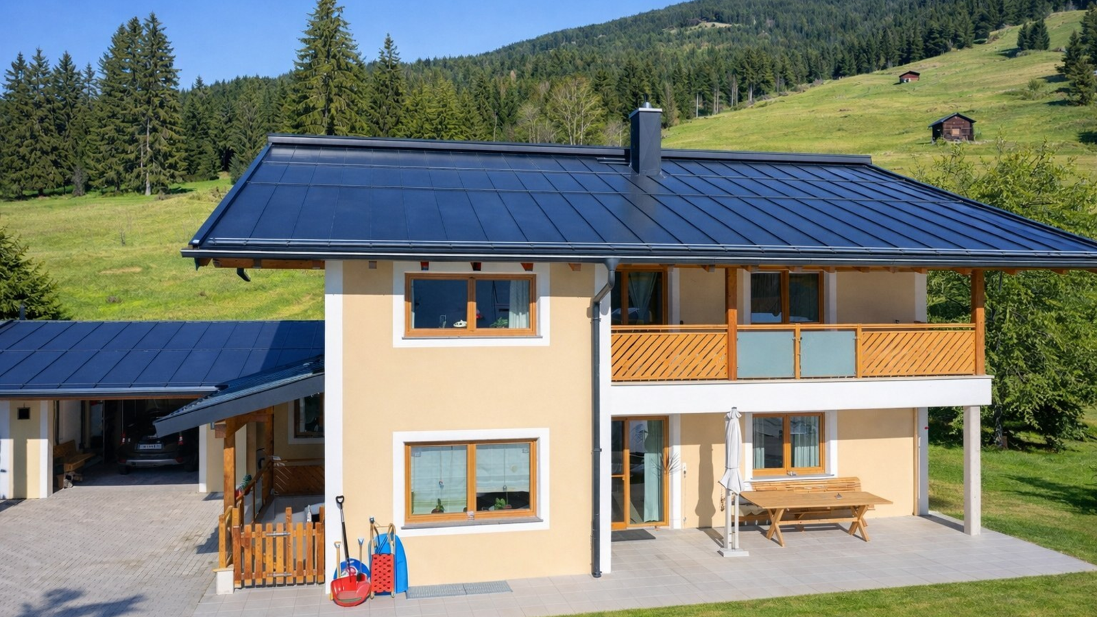 Exterior view of the Harlandhof featuring a balcony, terrace, and an attached carport against a forested mountain landscape.