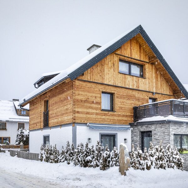 The winter exterior of the Farm House, showcasing a wooden upper facade, white lower level, and a stone-clad extension with a balcony.