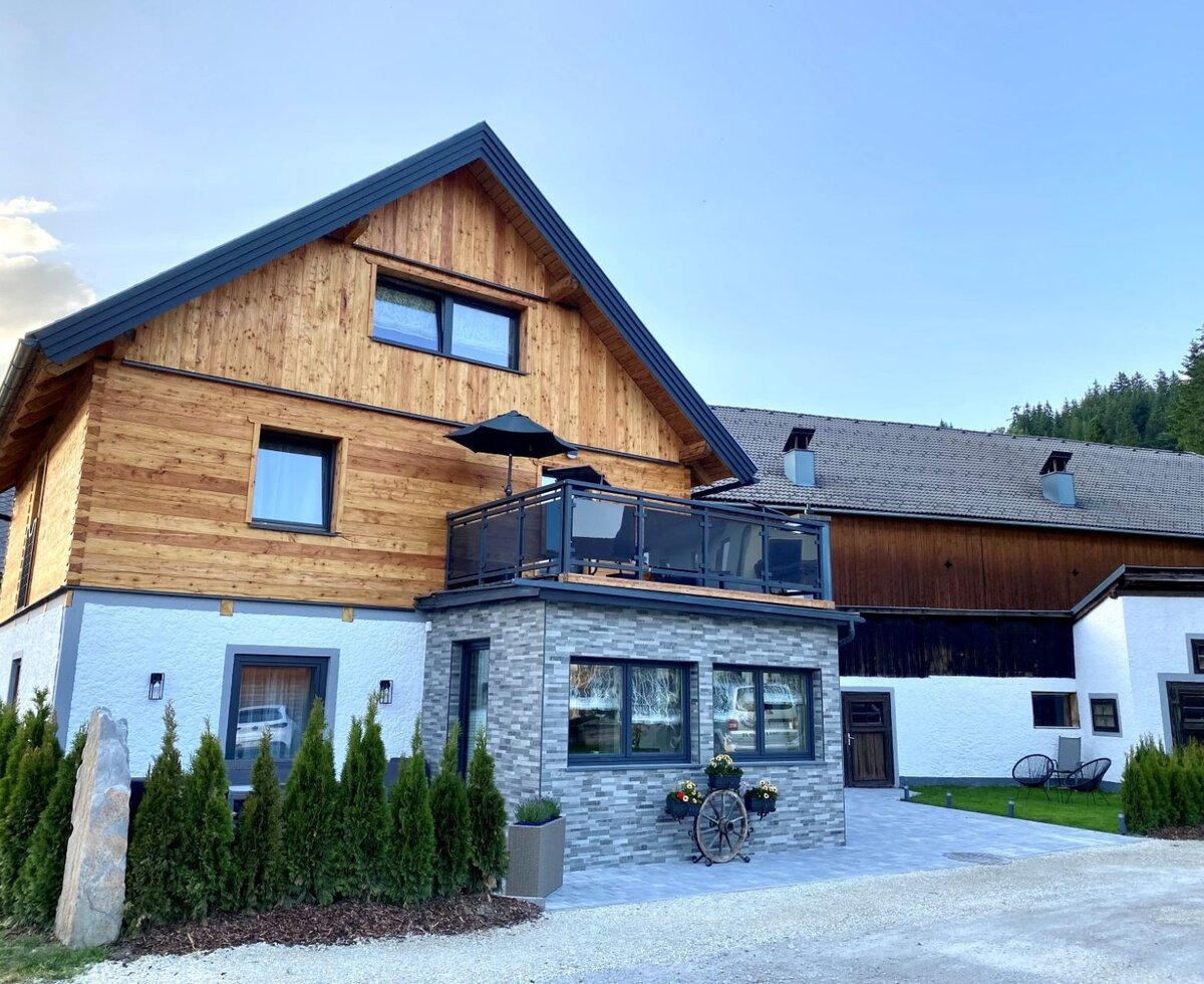 The farm house, featuring a wooden upper facade, a white lower level with a stone-clad extension, and a balcony, next to an adjacent barn-style building.