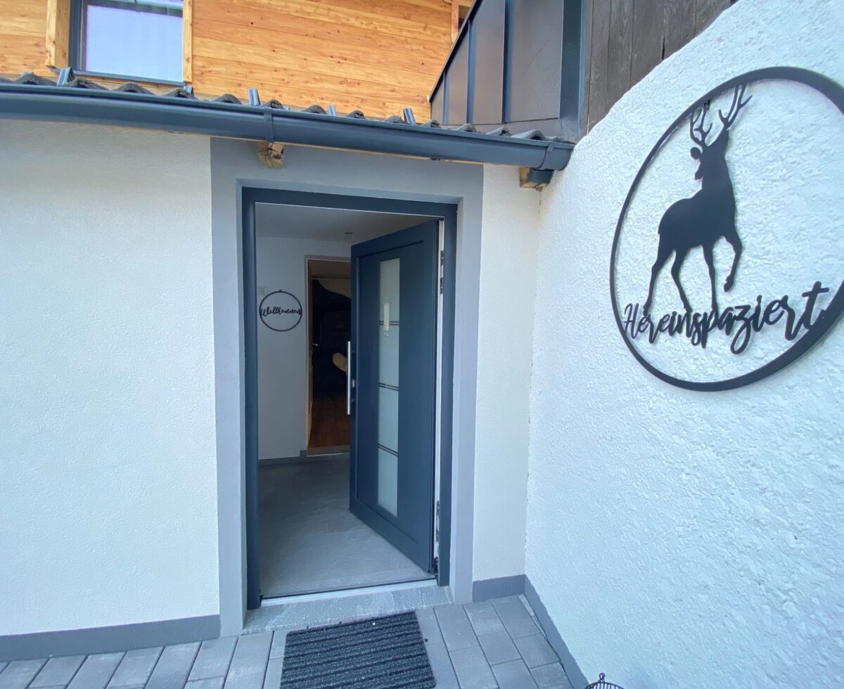 The main entrance to the farm house, featuring a grey door and a metal sign on the wall with a deer silhouette and the word "Hereinspaziert".