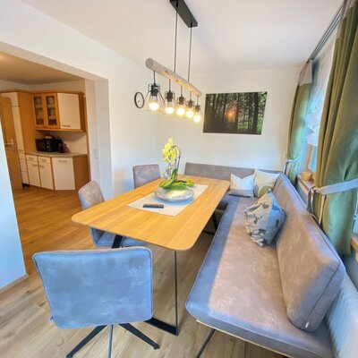 Dining area in the farm house with a wooden table, corner bench seating, additional chairs, and an overhead light fixture, with an adjoining kitchen visible.