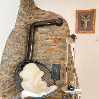 A corner of the farm house featuring a wood-burning stove set against a stone wall, with a black chair, a sheepskin throw, and a floor lamp.