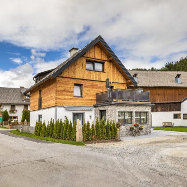 Exterior view of the farmhouse with wooden facade, balcony, and gravel parking area.