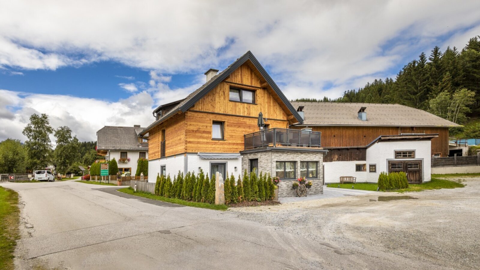 Exterior view of the farmhouse with wooden facade, balcony, and gravel parking area.