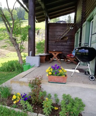 The covered porch of the farmhouse provides wooden outdoor seating and a grill, surrounded by garden plantings.