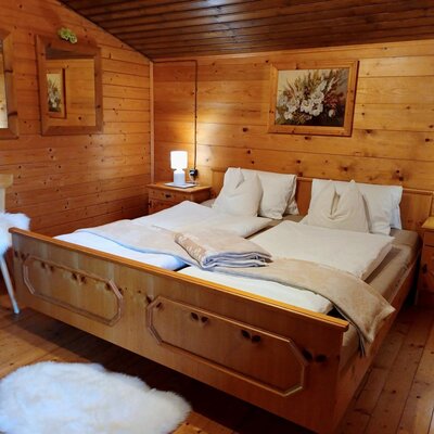 Double bedroom in the farmhouse with wooden walls and ceiling, a wooden bed, two bedside tables with lamps, and a wooden chair.