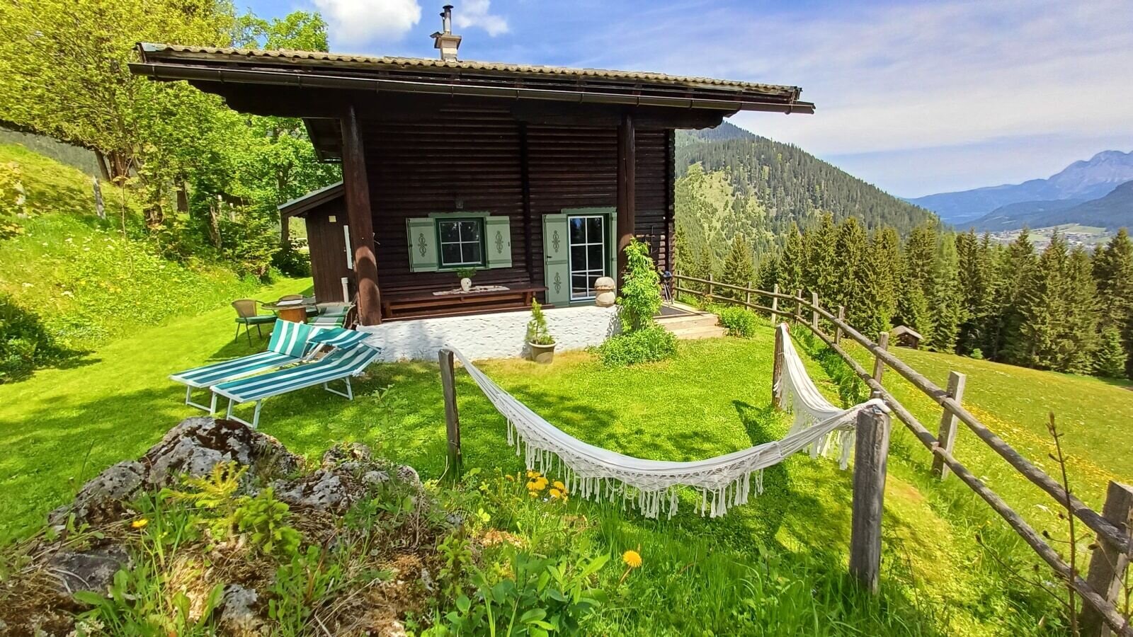 The farmhouse exterior, featuring a terrace with sun loungers and a hammock, overlooks a grassy hillside with mountain and forest views.