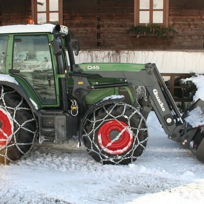 A green tractor equipped with snow chains parked in front of the farmhouse's rustic wooden building in winter.
