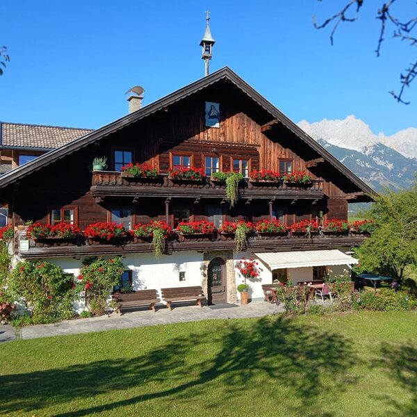 The inviting Hinterburghof farmhouse featuring flower-adorned wooden balconies and a sunny lawn against a scenic mountain backdrop.
