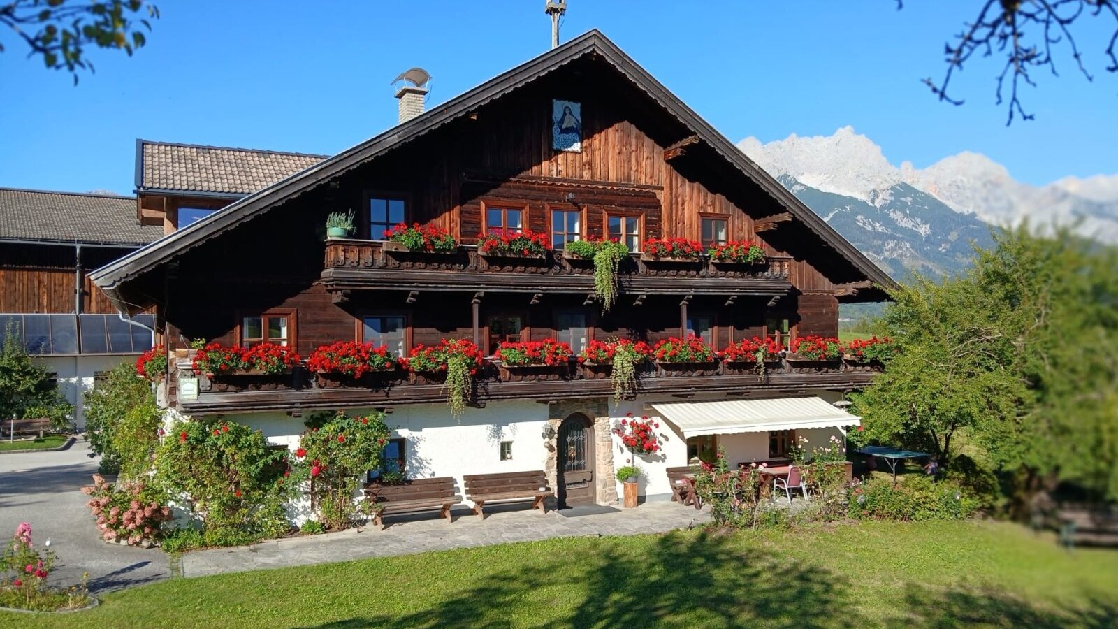 The inviting Hinterburghof farmhouse featuring flower-adorned wooden balconies and a sunny lawn against a scenic mountain backdrop.