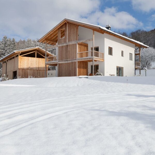 Exterior view of the farmhouse with a white facade, wooden cladding, and balconies, surrounded by a snow-covered landscape and forest.