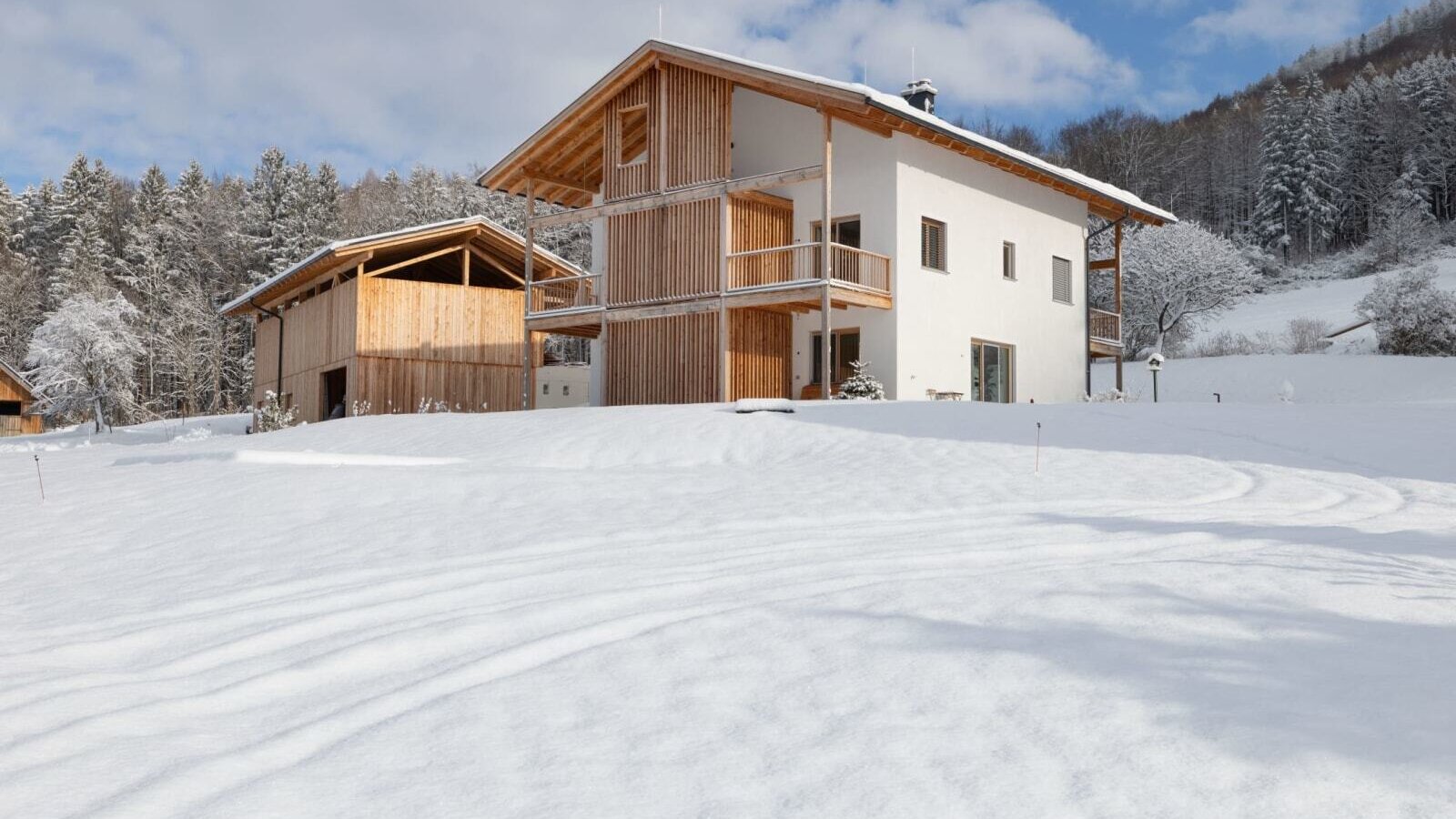Exterior view of the farmhouse with a white facade, wooden cladding, and balconies, surrounded by a snow-covered landscape and forest.