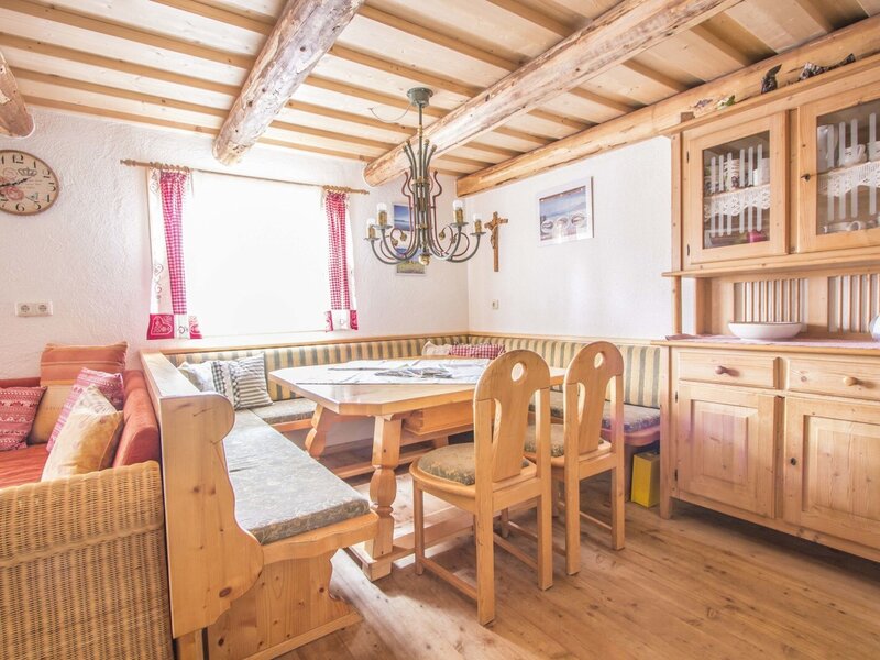 The dining and common area in the farmhouse features a wooden table, corner bench seating, additional chairs, a wicker sofa, and a wooden hutch, all under exposed wooden ceiling beams.