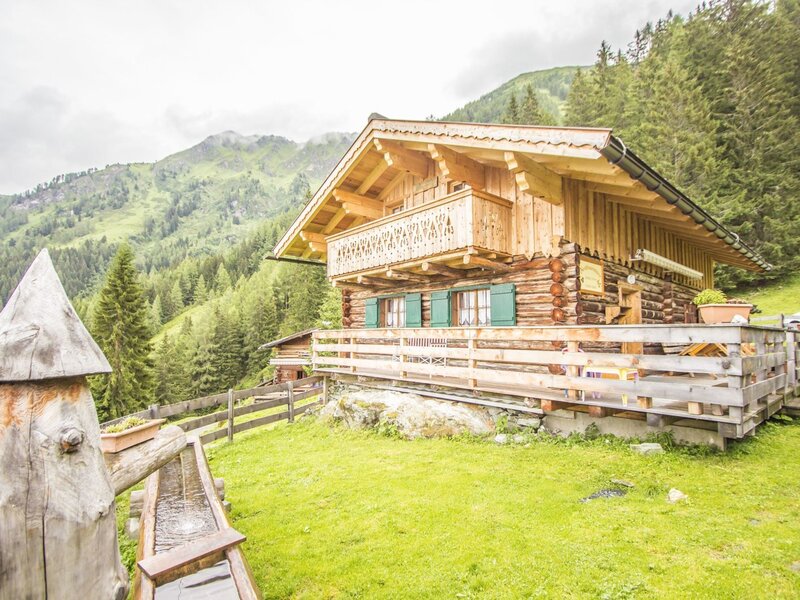 The wooden farmhouse exterior features a second-floor balcony, green-shuttered windows, a ground-level fenced terrace, and a wooden water feature in the foreground, set amidst mountains and forests.