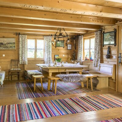 The cozy dining area of the alpine hut, featuring an inviting wooden table, rustic benches with warm fur throws, and colorful woven rugs.