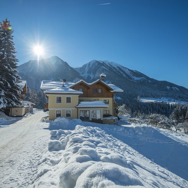 The exterior of the Farm House, a yellow building with wooden upper levels, nestled in a snow-covered mountain landscape under a bright sky.