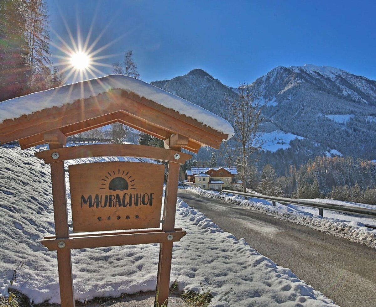 The inviting wooden sign of the Maurachhof farmhouse set against a sunny, snow-covered winter landscape with views of the Alps.