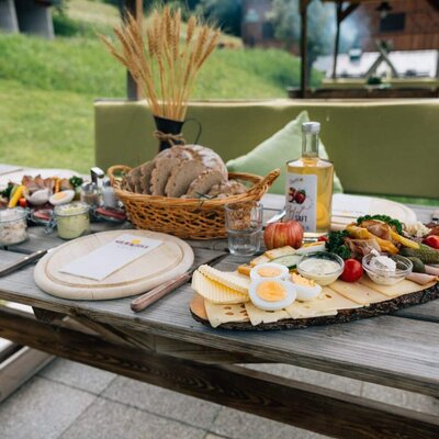 A wooden table at the farmhouse set with a rich, traditional platter of local cold cuts, cheeses, fresh bread, and juice in an outdoor setting.