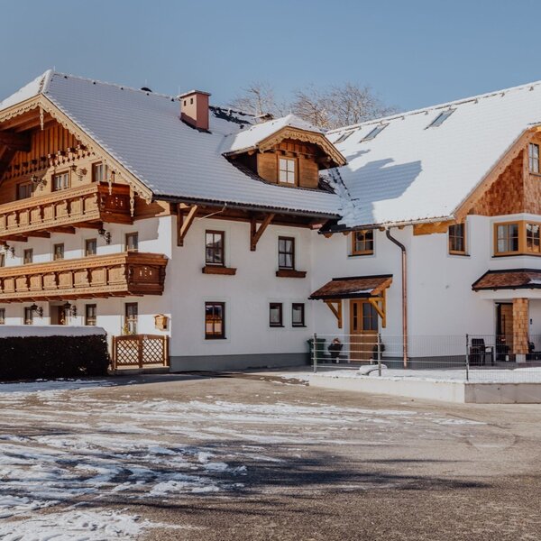 The farmhouse in winter, featuring a snowy roof and wooden balconies against a mountain backdrop.