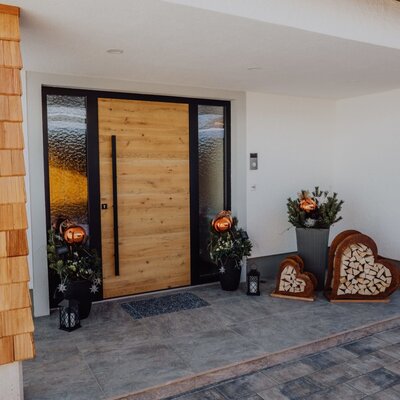 The entrance area of the farmhouse, featuring a wooden door, shingle-clad pillars, and decorations with plants and heart-shaped firewood holders.