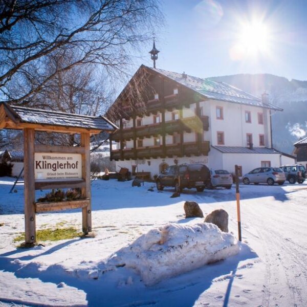 The Farm House exterior in winter, showing the welcome sign, parking area, and snow-covered surroundings with mountains in the background.
