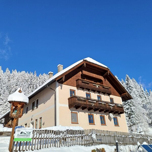 The Krametergut Farm House exterior in winter, featuring wooden balconies and a snow-covered forest background.