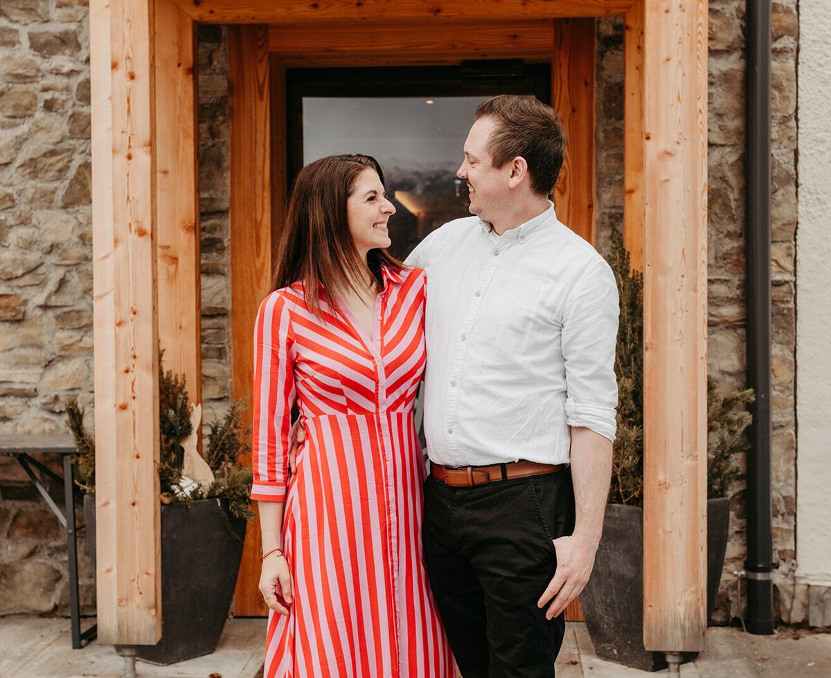 A couple standing in front of the entrance of the farmhouse with a wooden door frame and stone walls.